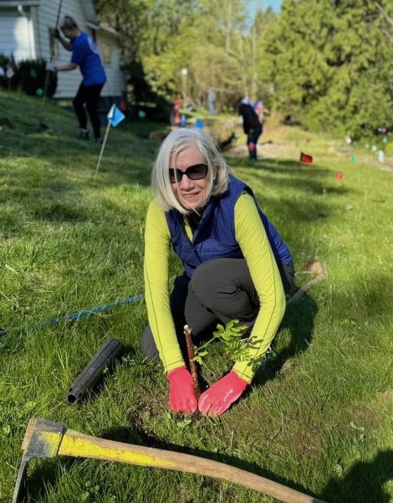 woman in sunglasses and a blue vest plants a plant, with a pick axe in front of her and other workers behind her