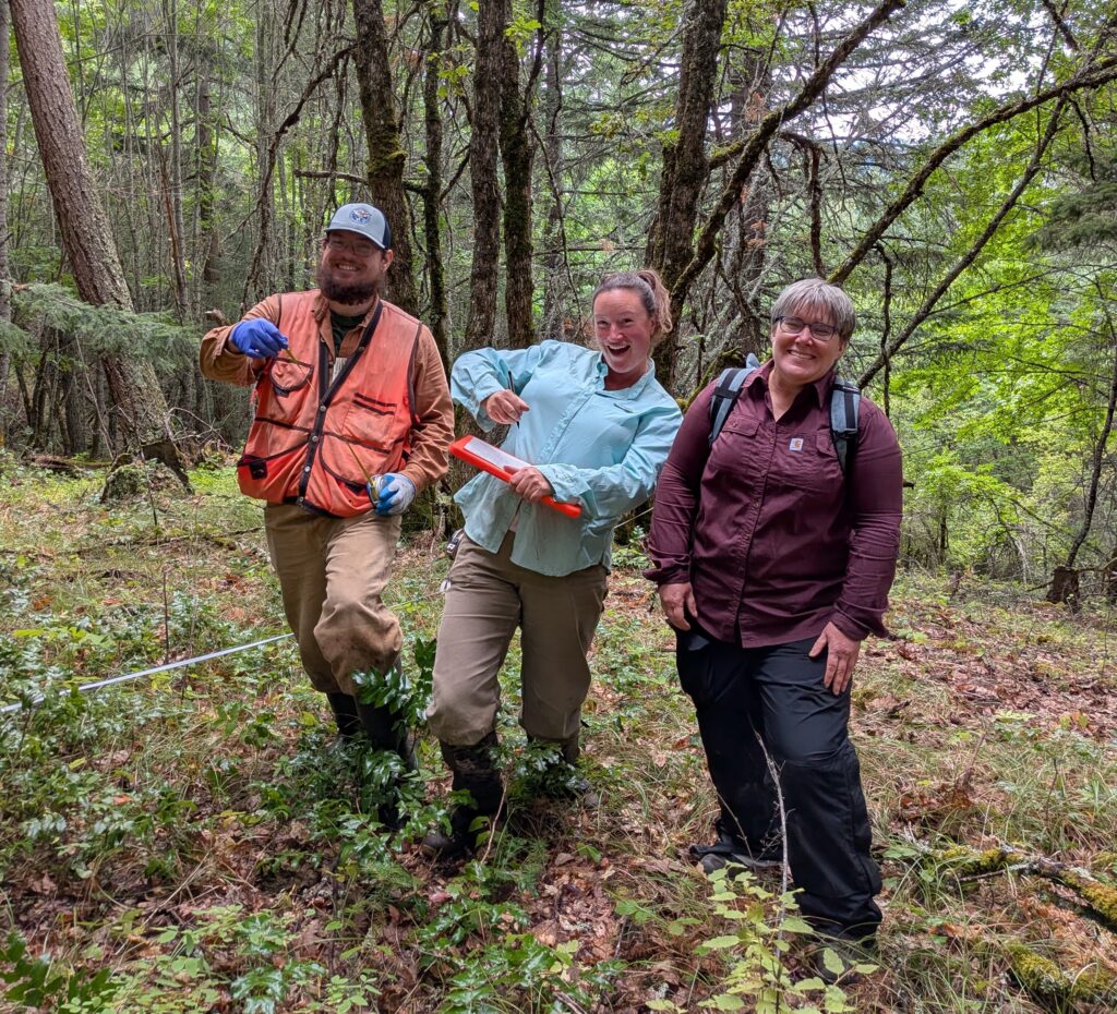 three smiling people stand together in the forest gesturing around them with their hands