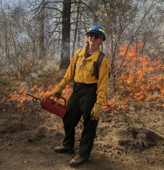 kat smiles standing in front of a prescribed fire holding a drip torch and wearing safety gear