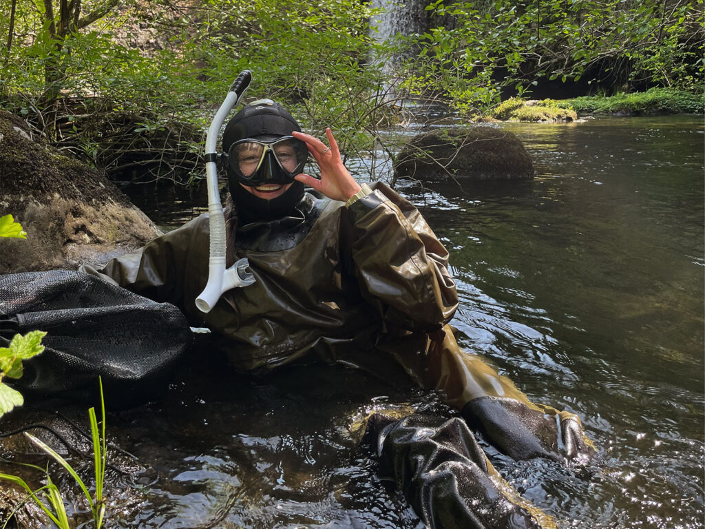 a person wearing a snorkel and a dry suit sits in waist deep water in a creek