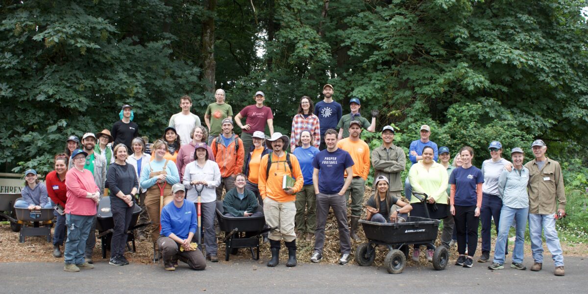 about 30 land trust staff and volunteers stand together in front of a small forest and piles of mulch