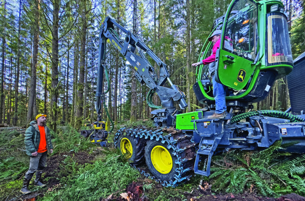 man in high visibility orange shirt stands next to a large piece of forestry equipment and talks to the driver
